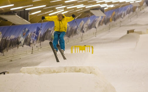 man met gele jas en blauwe broek maakt een ski sprong op de piste
