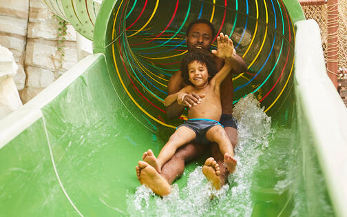 Blij kind dat met zijn vader languit op de rug van de grote groene waterglijbaan glijdt en de vader zwaait naar de camera