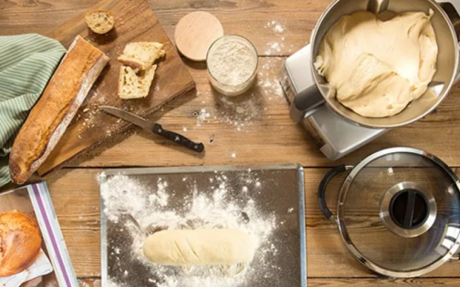 houten tafel met keukengerei: snijplank met stokbrood, keukenrobot, en gerief om brood te bakken