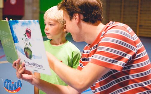 lesgever met kind kijkend in boek 'de mondenboom' met onderaan logo van MiLa