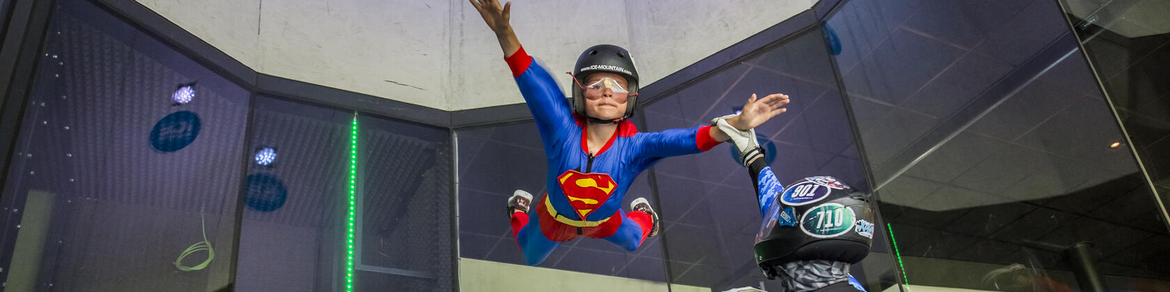 vrouw met superwoman kostuum is aan het indoor skydiven, een begeleider staat er naast om te helpen waar nodig